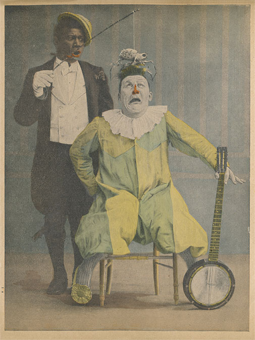 Photo of Chocolat the Clown wearing a vest and tailcoat and a yellow hat. He is standing with a comical look on his face as he dangles a fake spider over Foottit’s head. Foottit is sitting, holding a banjo and wearing a yellow clown costume. He looks scared.