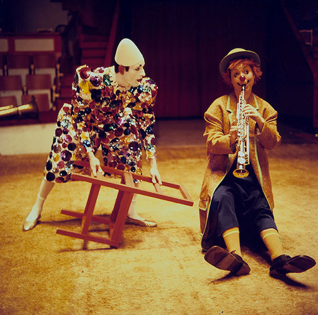 Photo of a fair-skinned woman with red hair and wearing a red nose and oversized shoes. She is sitting in the middle of a ring, playing saxophone. Next to her, a man in a sequined clown costume is holding a red chair. He is wearing a pointy white hat. He has white makeup on his face and red makeup on his ears.