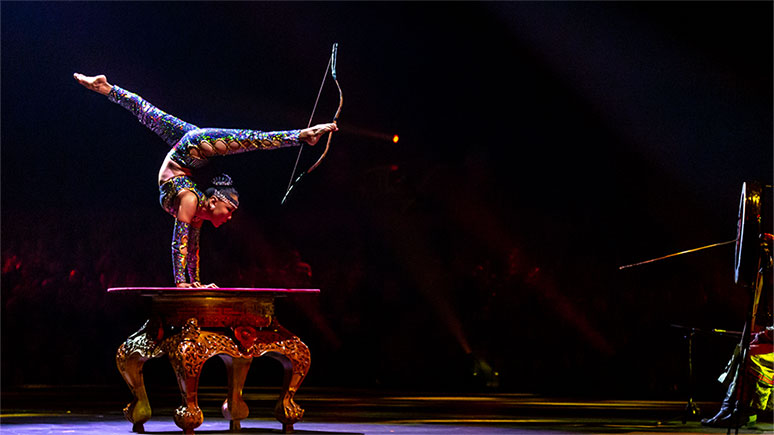 Photo of show where a woman in a blue sequined costume does a handstand on a large table. She has her legs over her head. She is holding a bow with her right foot and appears to have shot an arrow with her left.