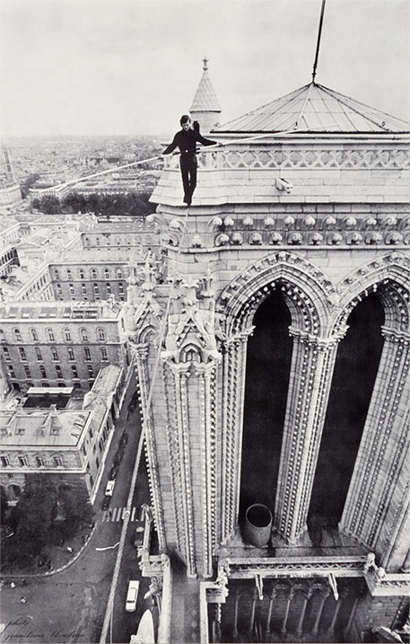 Black and white photo from a high vantage point of a man balancing on a wire. The vehicles far below look like toys.