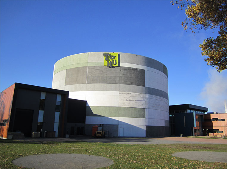 Photo du site de la TOHU. Au centre, le bâtiment principal est cylindrique et coloré de différentes teintes de gris. Sur les côtés, les bâtiments bruns sont plus petits et plus ordinaires.