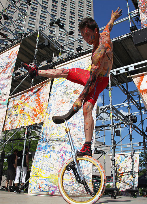 Photo of a fair-skinned bare-chested man who is covered in paint. He is balancing on one leg on the wheel of his unicycle.  He is performing in the street, with scaffolding in the background.