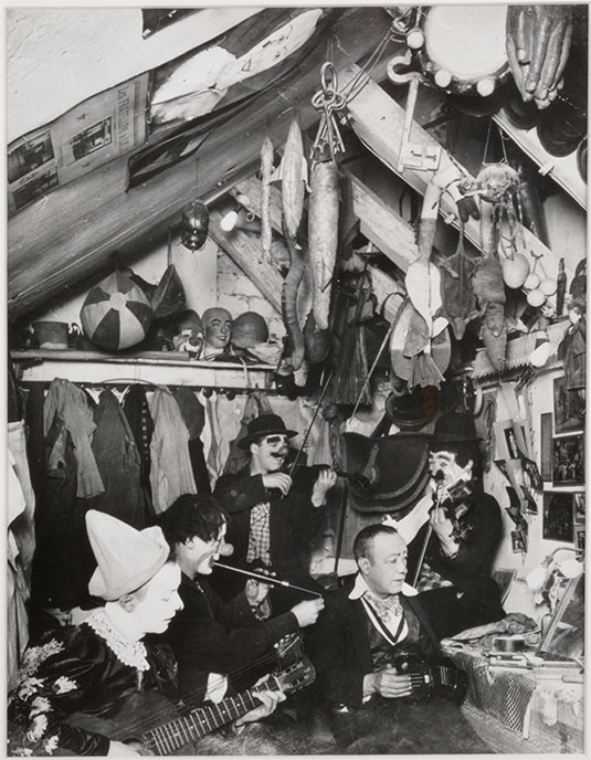 Photo en noir et blanc de cinq hommes jouant de la musique dans une petite pièce remplie d’accessoires en tout genre, du sol au plafond.